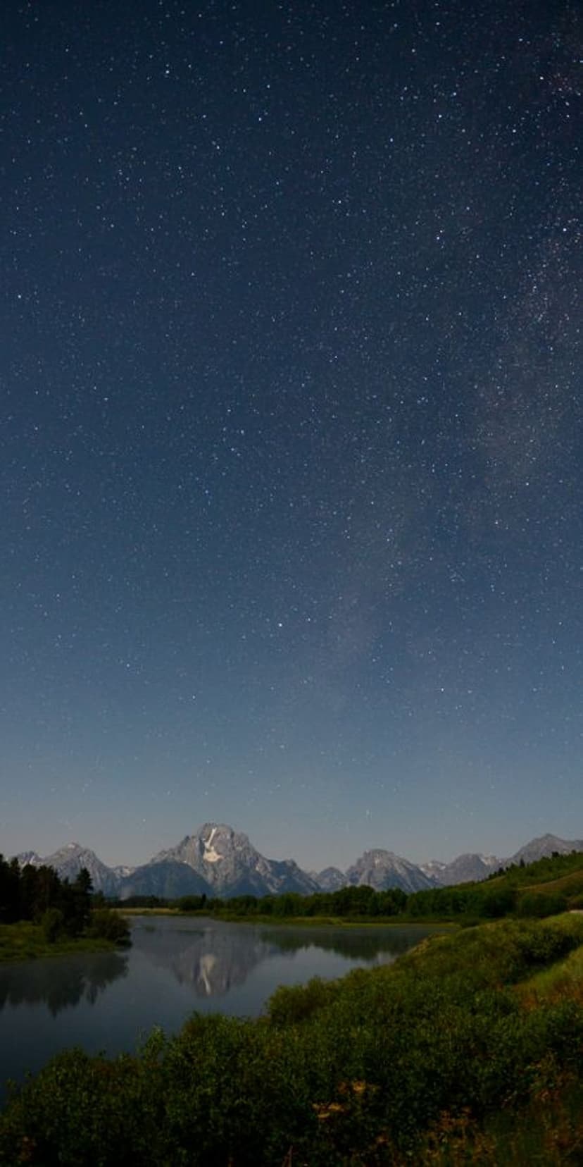 Rugged mountain peaks reflect in a dark lake beneath a dense canopy of stars and shimmering celestial clusters.