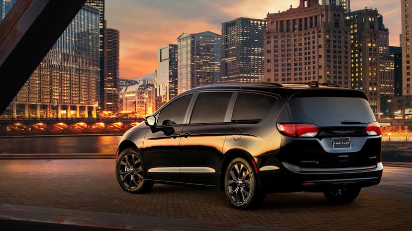 Black Chrysler Pacifica minivan parked on a brick pier with illuminated city skyscrapers during a golden hour sunset.