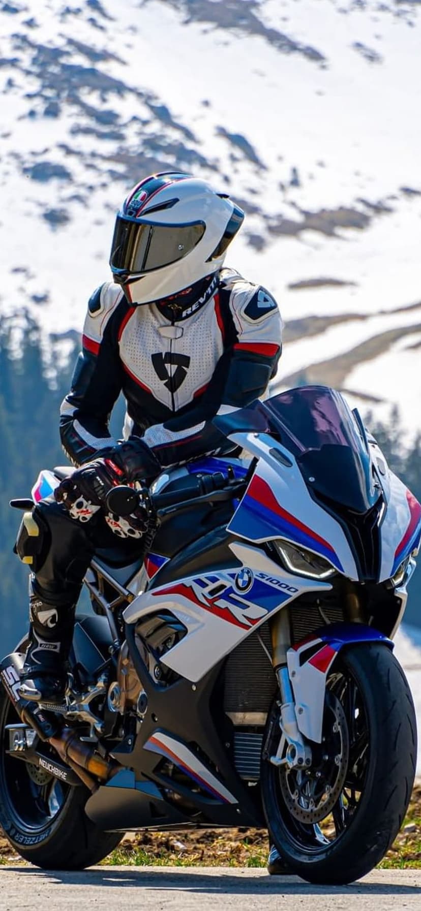 Motorcyclist in racing gear sits on a BMW S1000RR amidst a snow-covered mountain peak with distant evergreen trees.