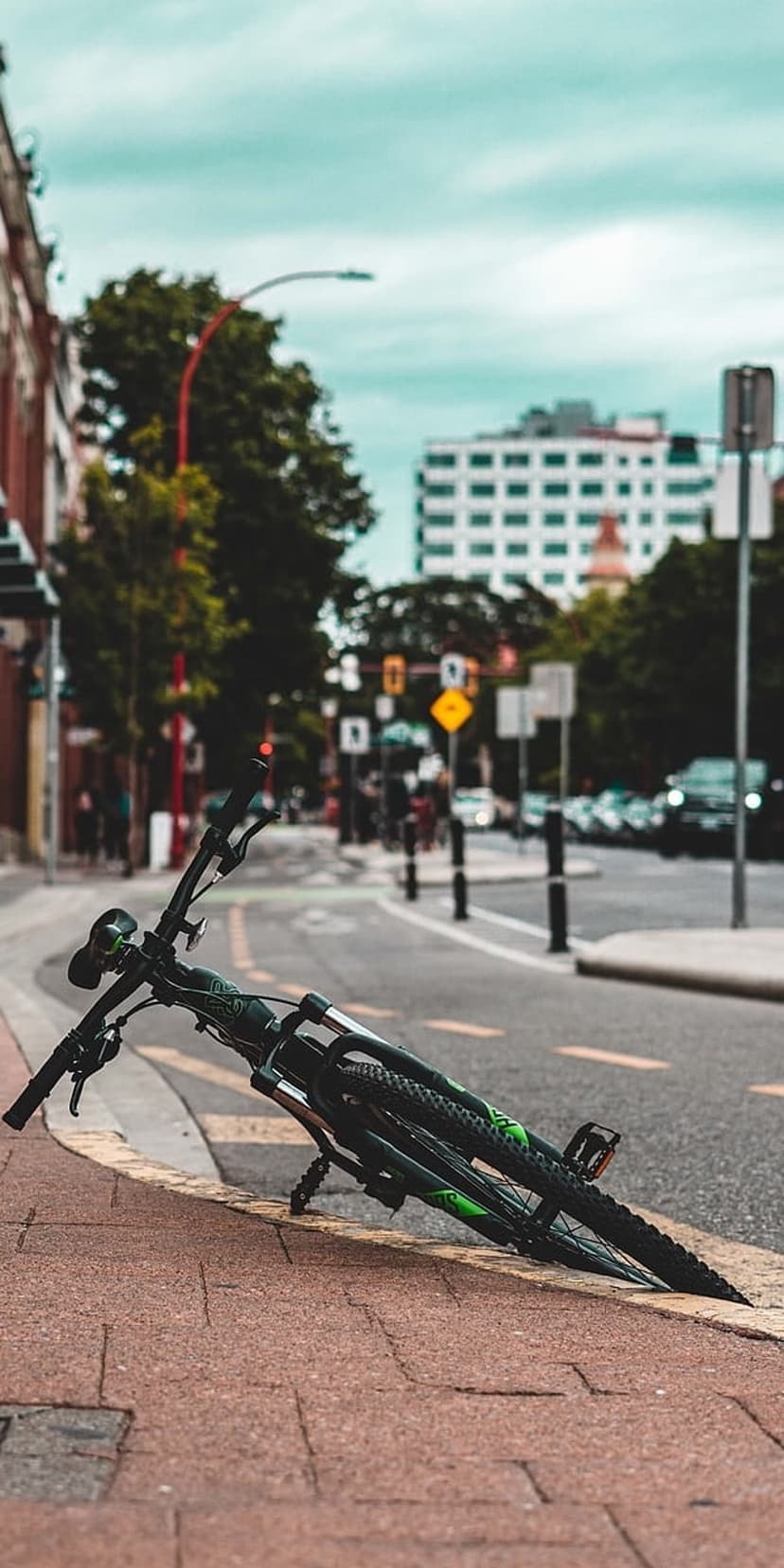 Black and green mountain bike lying on a red brick sidewalk against a teal cloudy sky and city street backdrop.