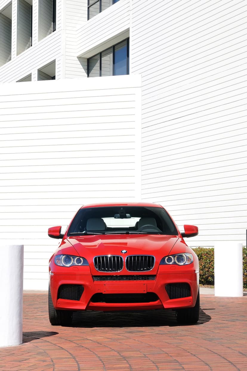 Front view of a red BMW X6 M parked on a brick path against a white modern building with horizontal siding.