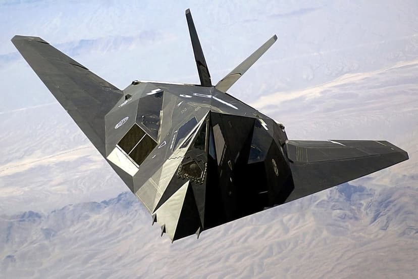 F-117 Nighthawk stealth fighter jet flying above brown desert mountains under a clear blue sky during the day.