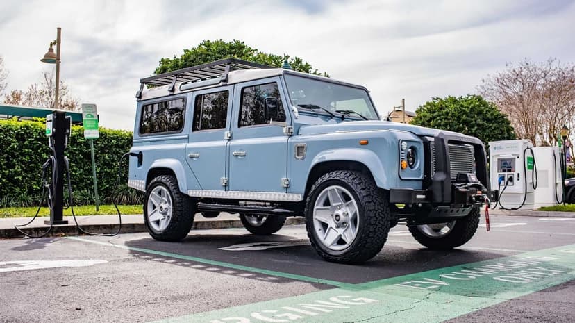 Light blue classic Land Rover Defender plugged into an EV charging station in a parking lot under soft daylight.