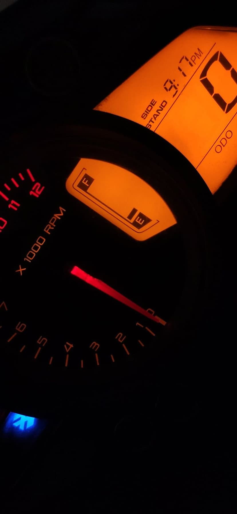 Motorcycle dashboard displays a glowing orange tachometer, digital clock, and fuel gauge in the dark night.
