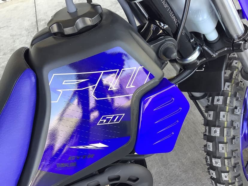 Close-up of a blue Yamaha PW50 fuel tank with white graphics and a rugged knobby off-road tire.