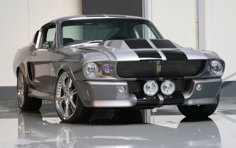 Metallic grey 1967 Shelby GT500 Mustang with black stripes and chrome wheels in a reflective studio setting.