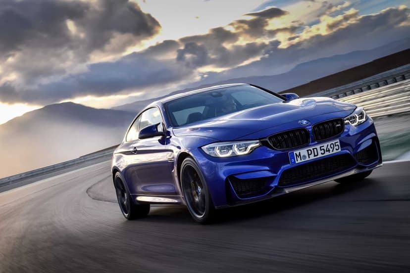 Blue BMW M4 sports car racing around a track curve with mountains and a sunset sky in the background.