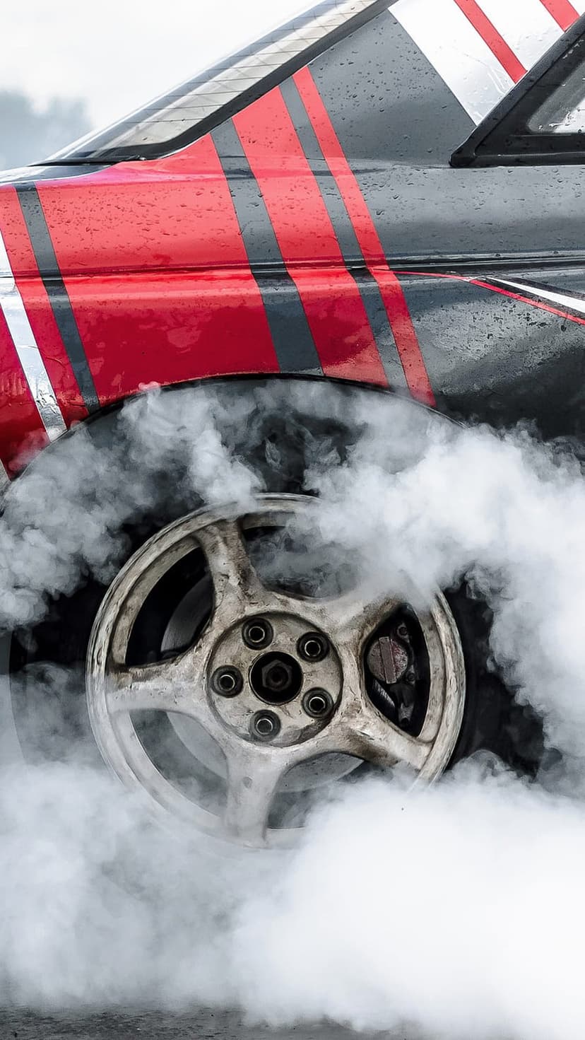 Race car wheel spinning amidst dense white smoke with red and black striped bodywork visible on a wet surface.