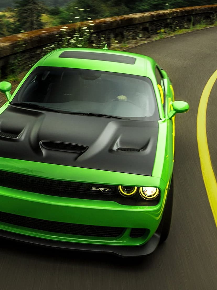 Green Dodge Challenger SRT with a black hood drives along a curved forest road with illuminated headlights.