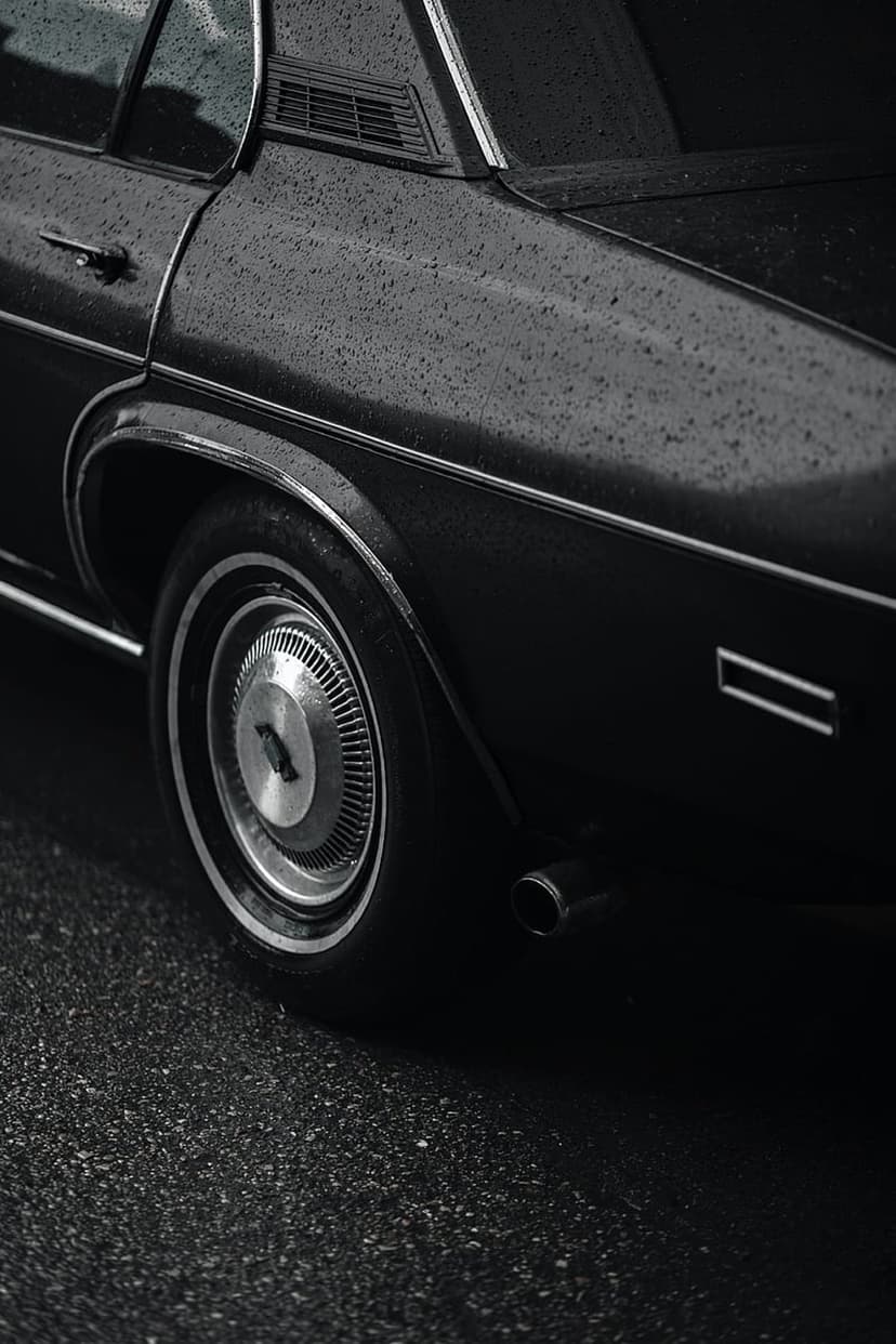 Vintage car rear fender with raindrops on black paint and a polished chrome hubcap on a wet asphalt road.