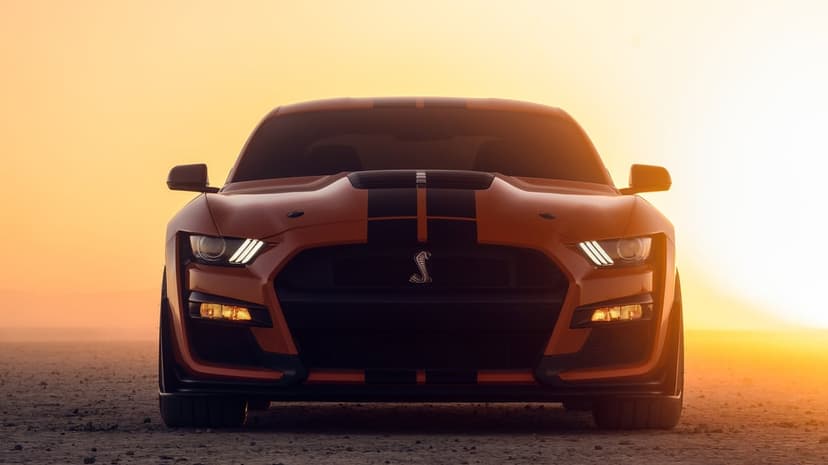 Orange Shelby GT500 Mustang with black stripes centered in a desert at sunset with bright LED headlights.
