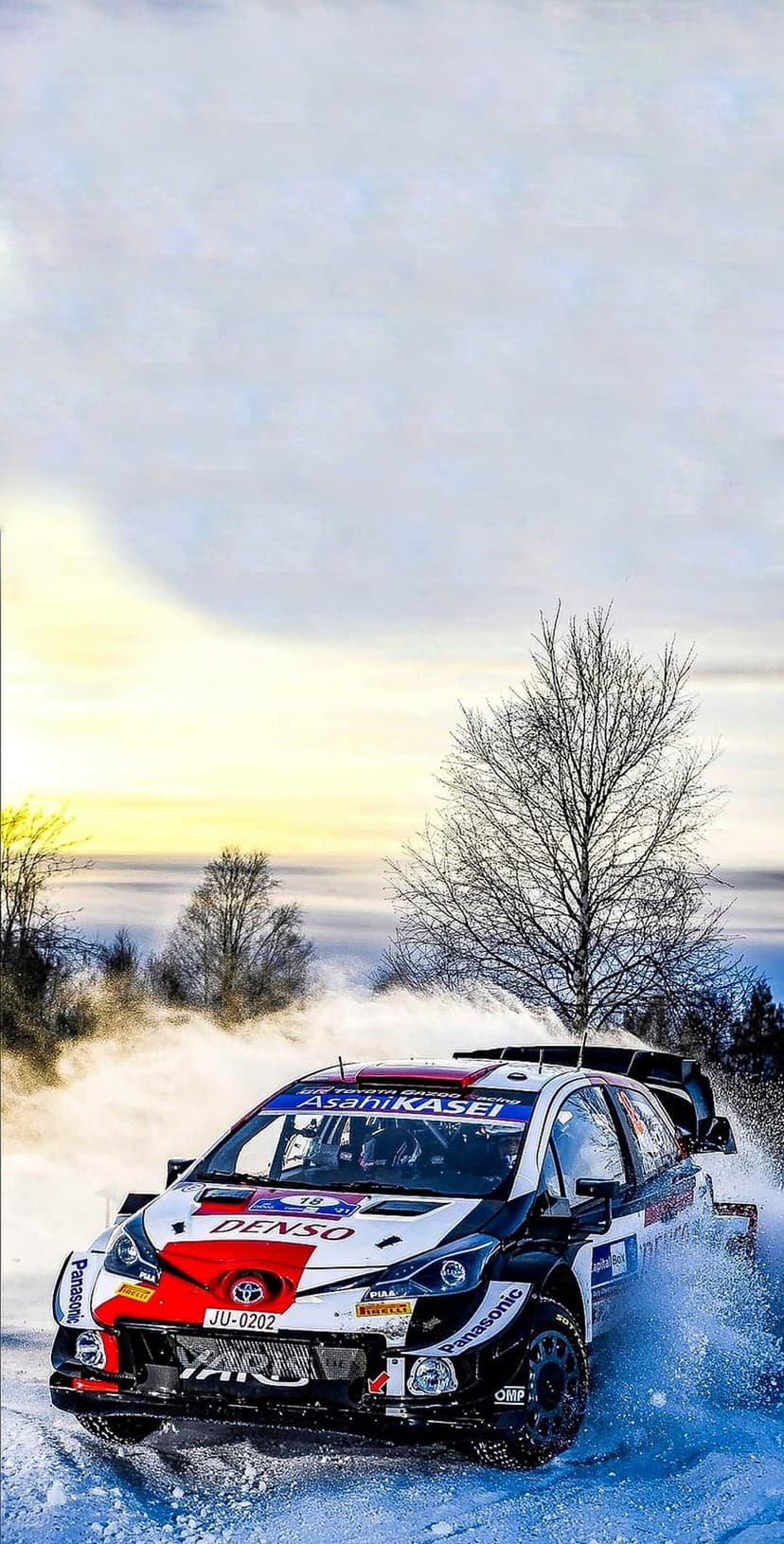 Toyota Yaris WRC car racing through deep snow on a winter track with bare trees under a bright hazy sky.