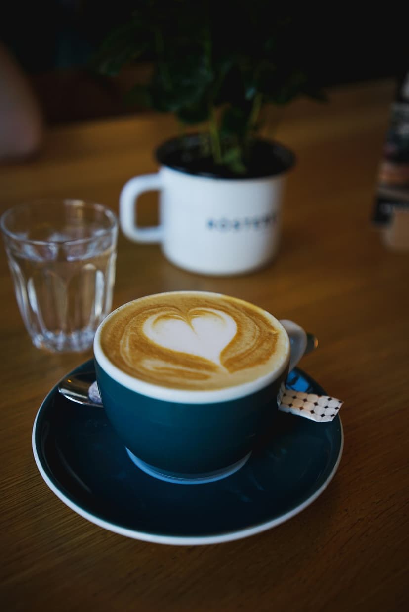 Heart-shaped latte art in a teal cup sits on a wooden table next to a water glass and a small potted plant.