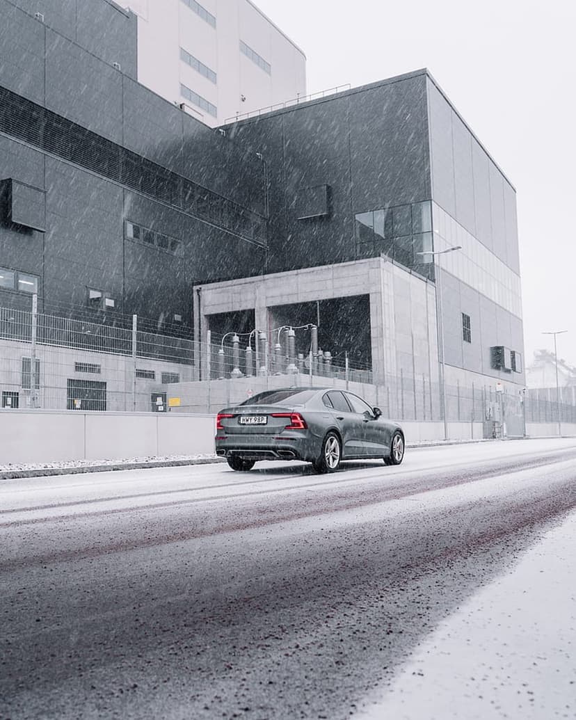 Grey sedan driving on a slushy road past a dark industrial building while snowflakes fall during winter.