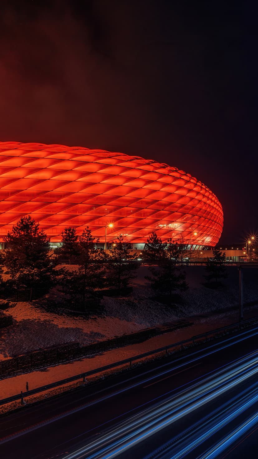 Large oval stadium glowing red under a dark cloudy sky with blue light trails from traffic on a lower road.