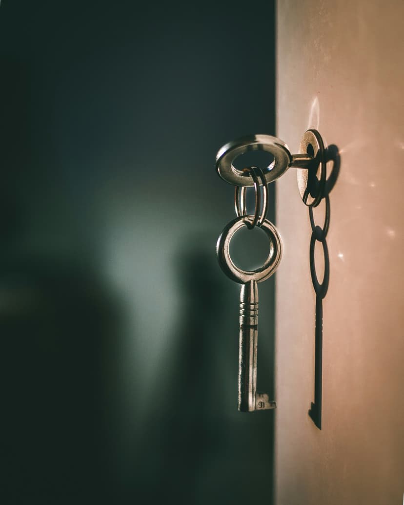 Two vintage metal keys, one ornate silver and one simple black, hang by a chain against a textured gray wall.