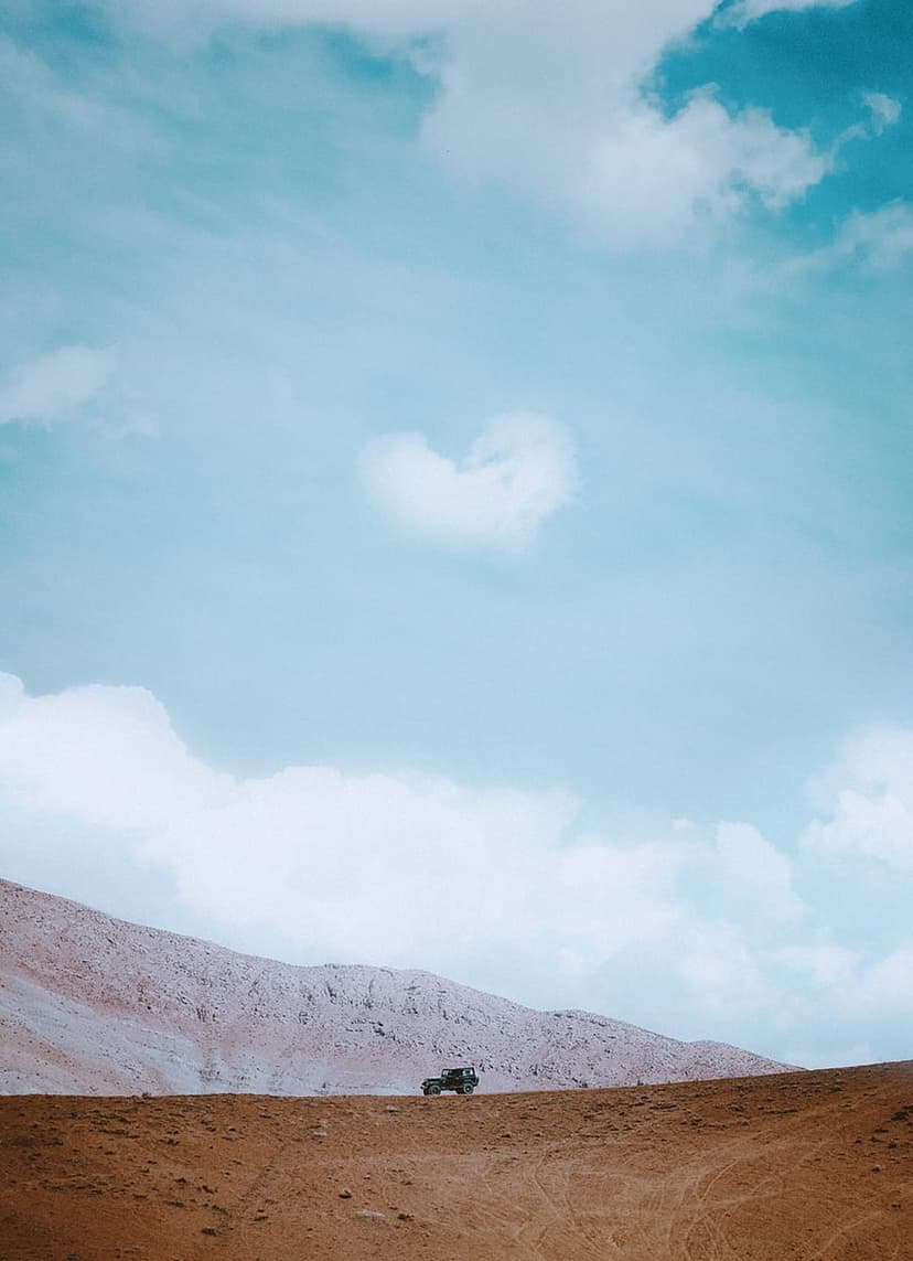 Rugged 4x4 truck navigating a dusty brown trail beneath a heart-shaped cloud and snow-capped mountain peaks.