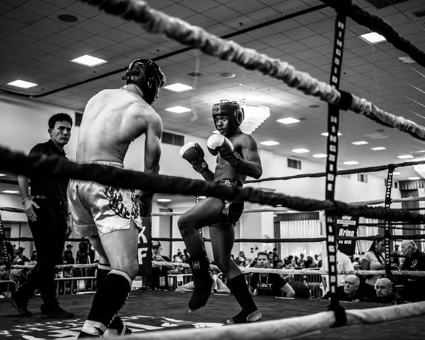 Muay Thai fighter lands a high kick on an opponent in a black and white boxing ring during a training session.