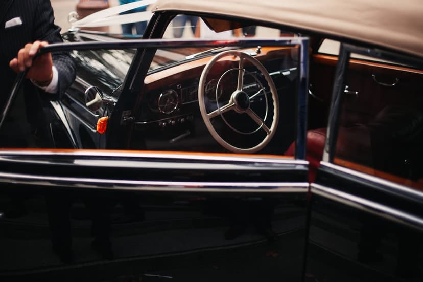 Man in a dark suit holding the door of a vintage car with a wooden dashboard and red leather upholstery.