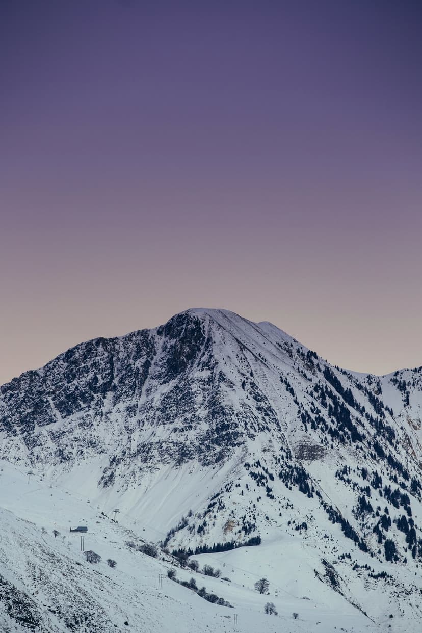 Snow-capped mountain peak with rocky ridges and evergreen trees under a purple and blue twilight gradient sky.