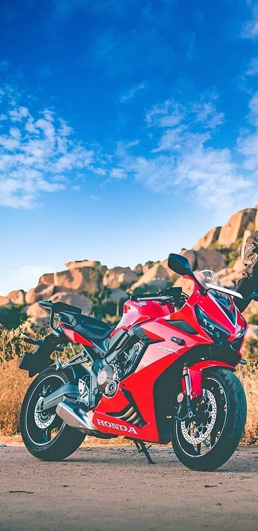 Red Honda CBR sportbike parked on a dirt mountain path with rocky peaks and a clear blue sky background.