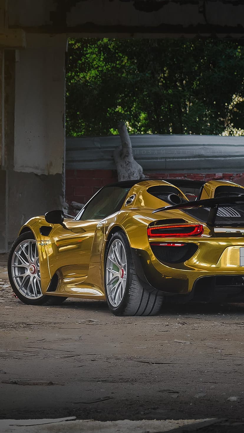 Porsche 918 Spyder with a gold wrap parked inside a concrete industrial building with green trees behind it.