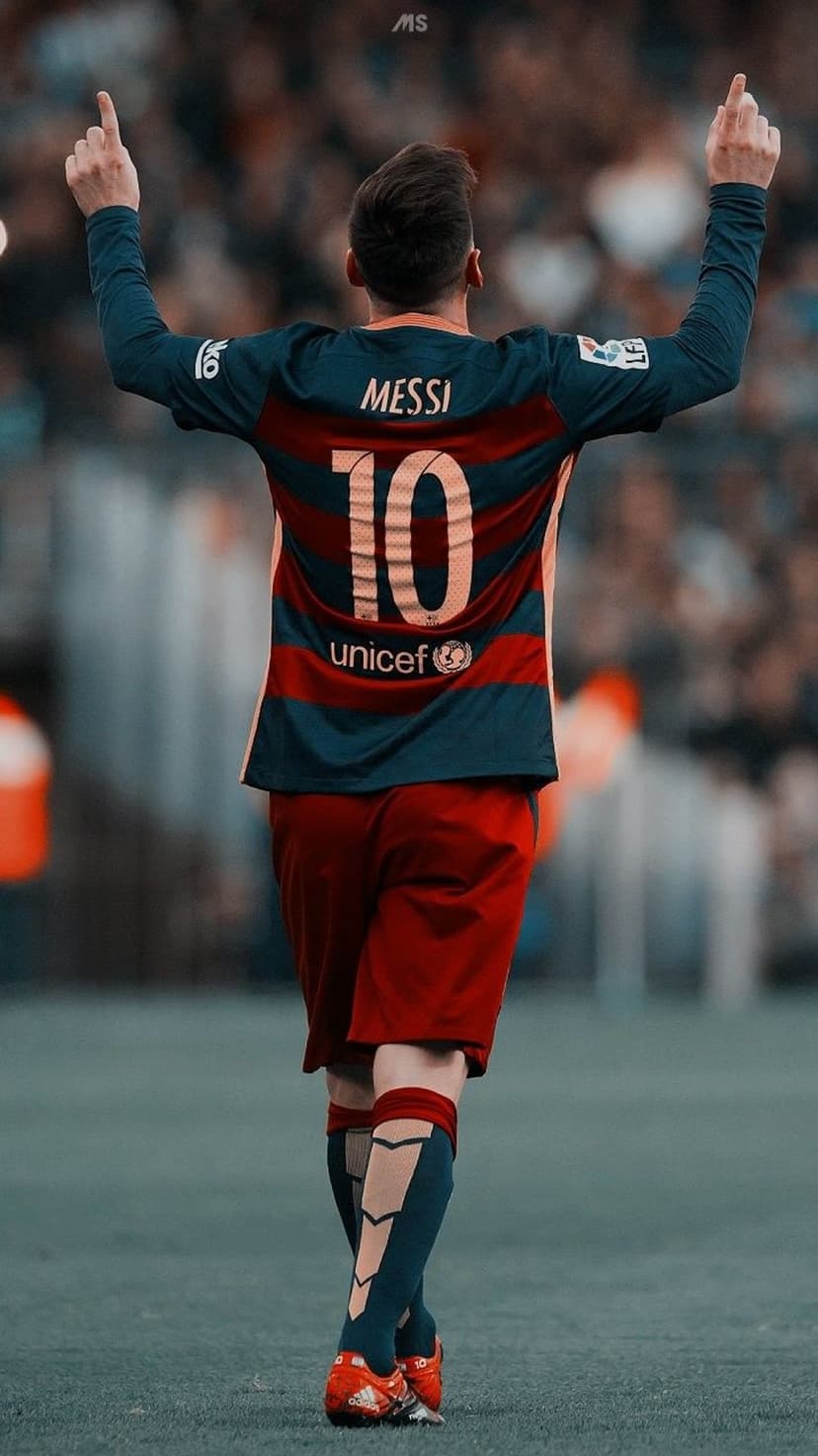 Lionel Messi stands on a football pitch wearing a Barcelona jersey with both arms raised pointing to the sky.