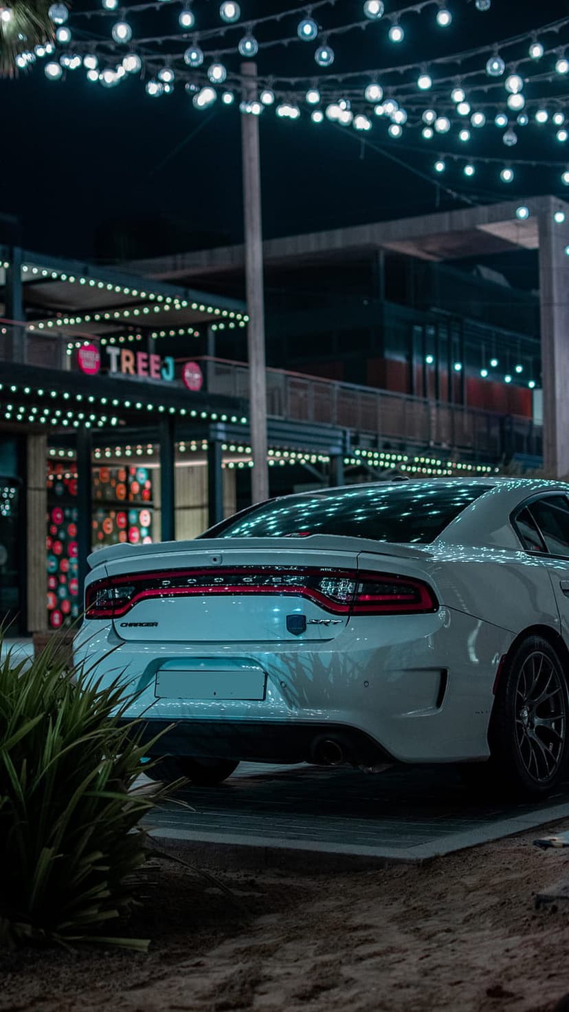 White Dodge Charger rear view parked in front of a building with glowing string lights and green leaves at night.