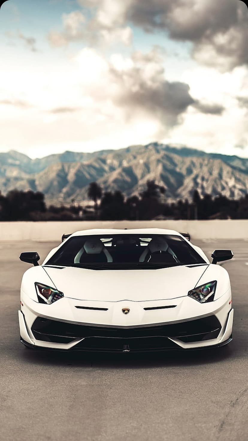 White Lamborghini Aventador S parked on asphalt with jagged mountains and a cloudy sky in the background.
