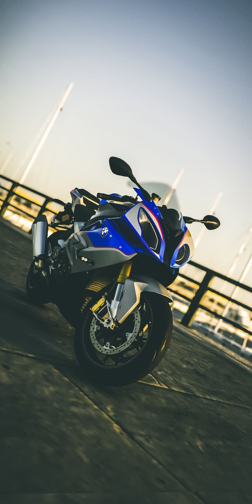 BMW S1000RR motorcycle in blue and white parked on a wooden dock with sunset lighting and boat masts behind it.