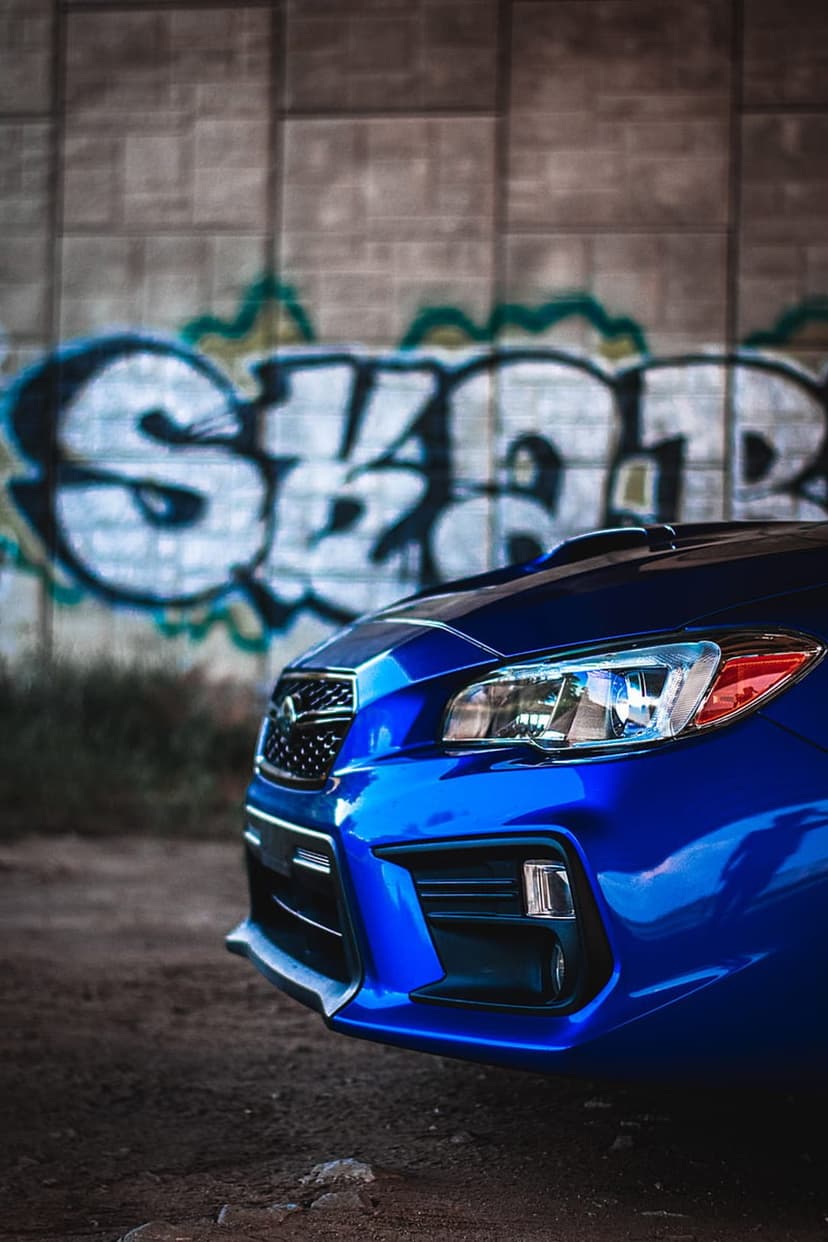Blue Subaru WRX front end sits before a concrete wall covered in vibrant multi-colored spray paint graffiti.