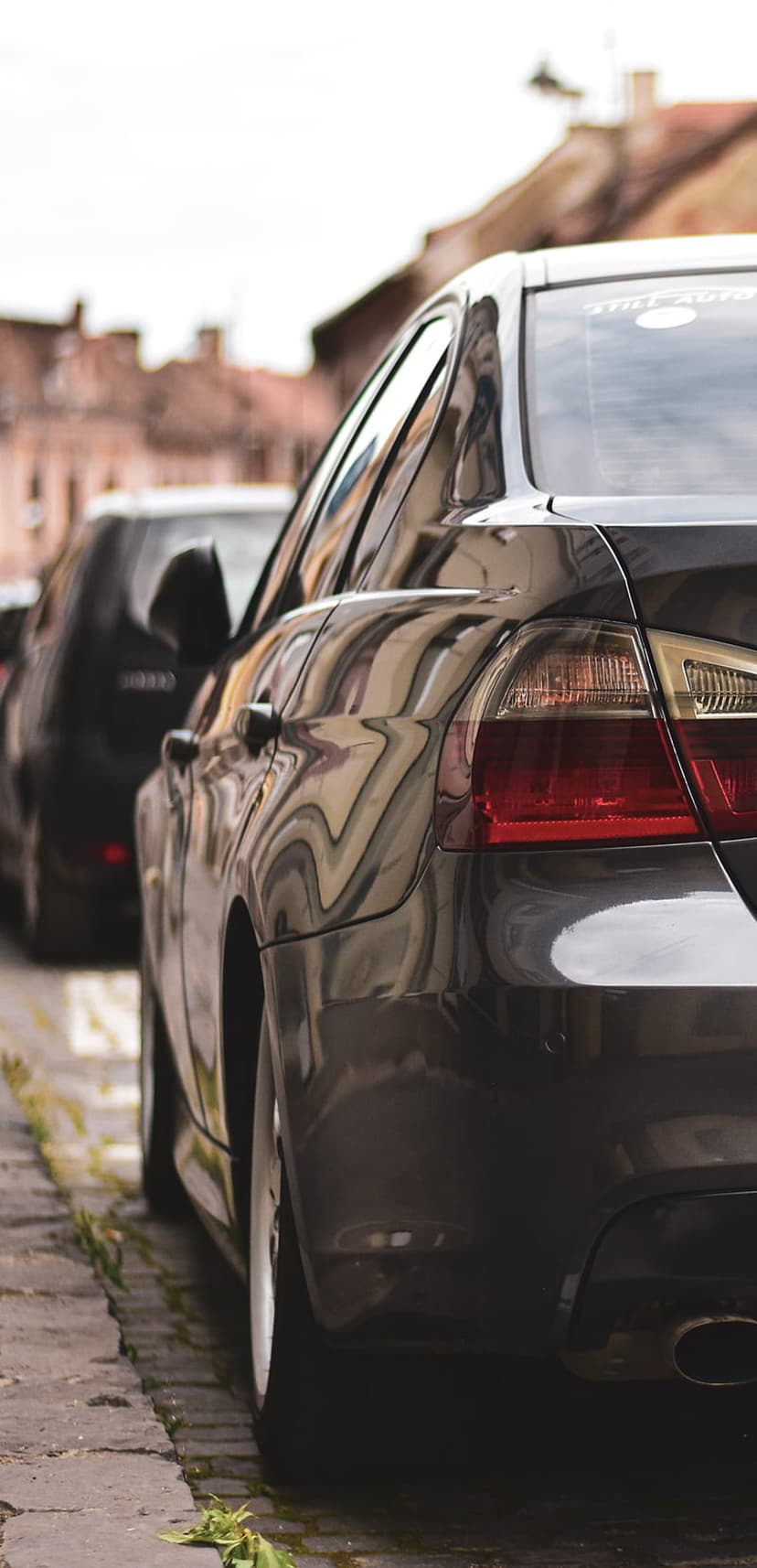 Rear taillight and polished fender of a dark sedan reflecting city buildings on a textured cobblestone street.