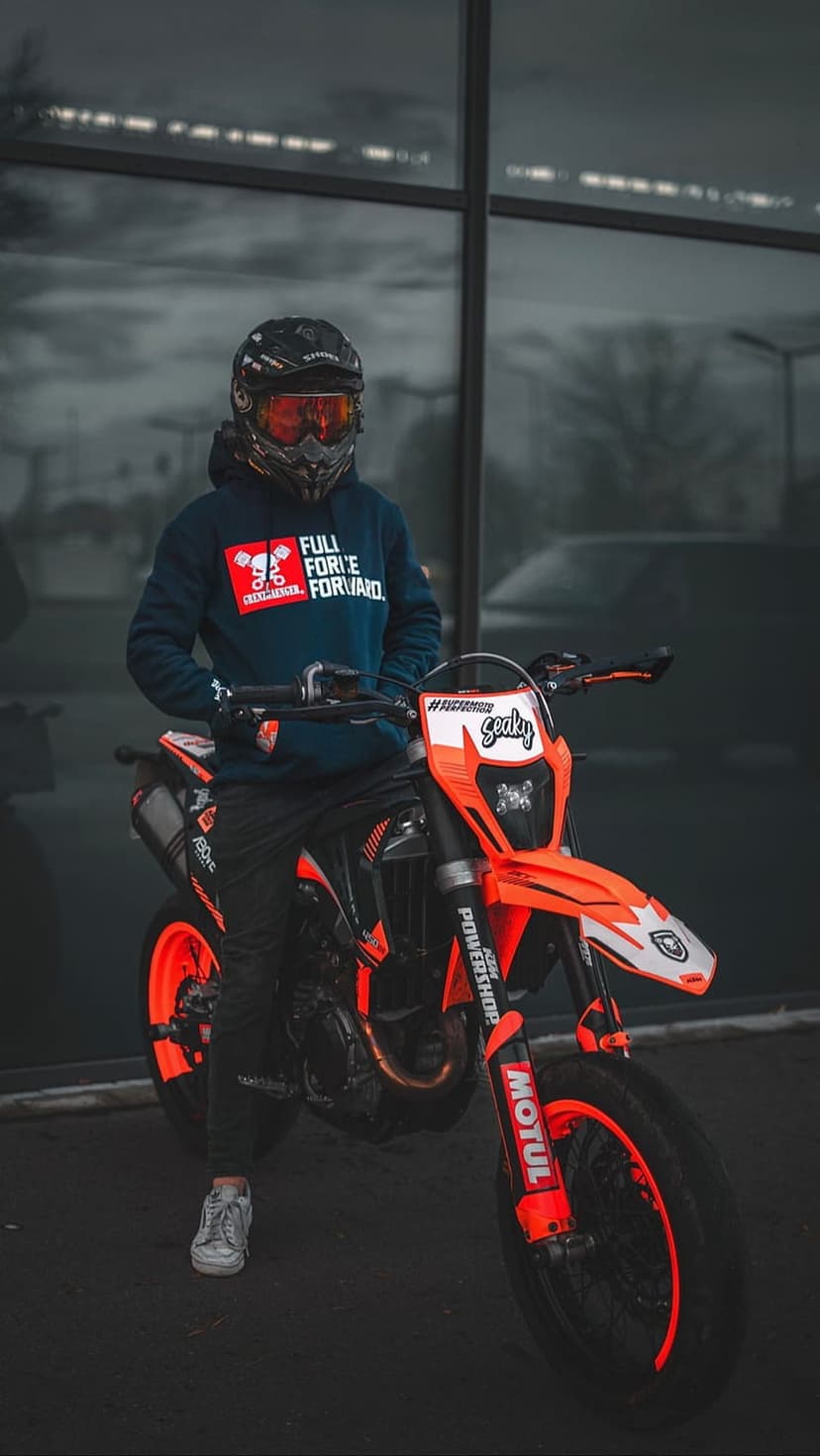 Rider in a helmet and blue hoodie standing by an orange supermoto motorcycle outside a modern glass building.