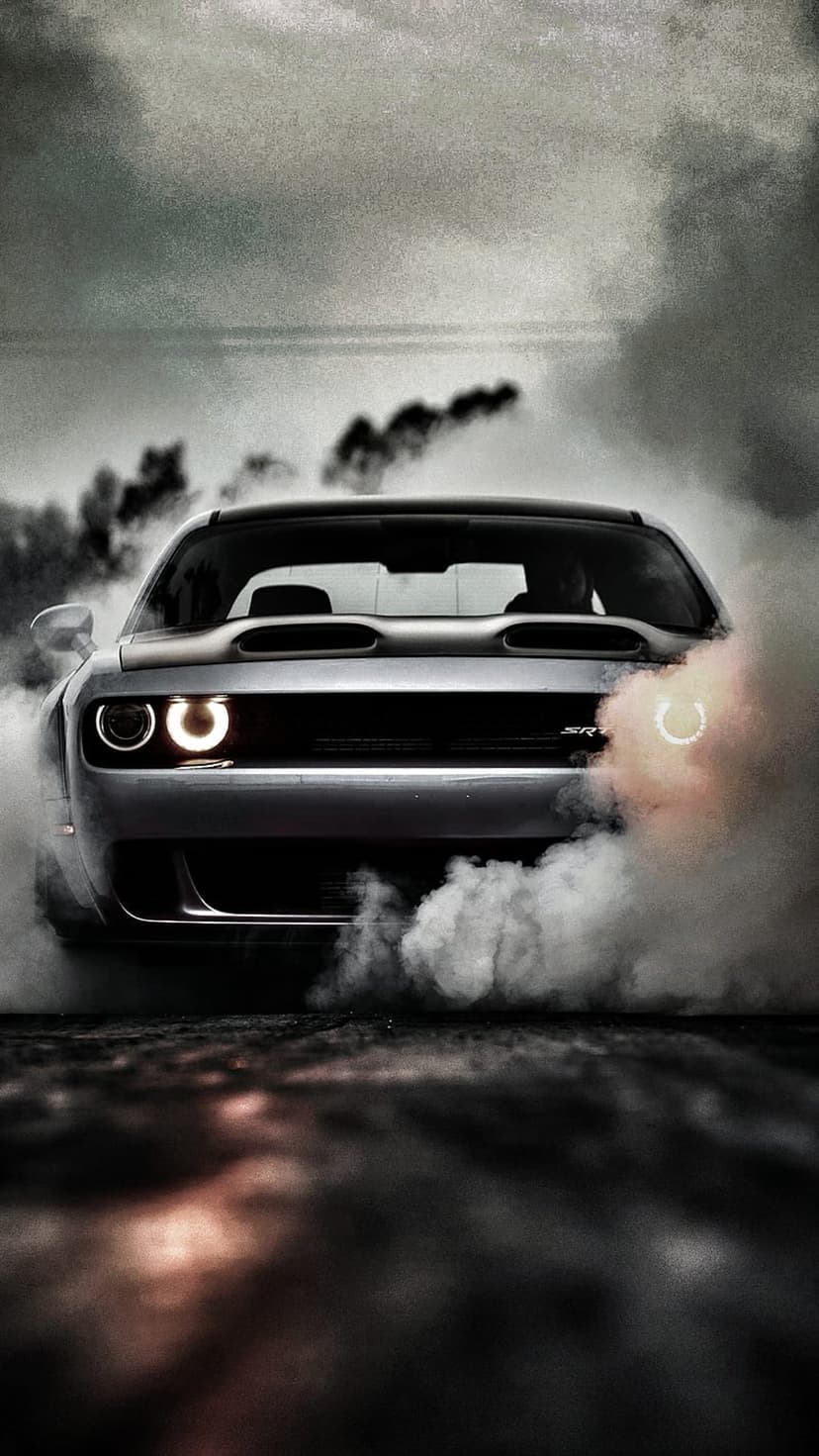 Silver Dodge Challenger SRT Hellcat performing a burnout with thick white smoke and glowing headlights.