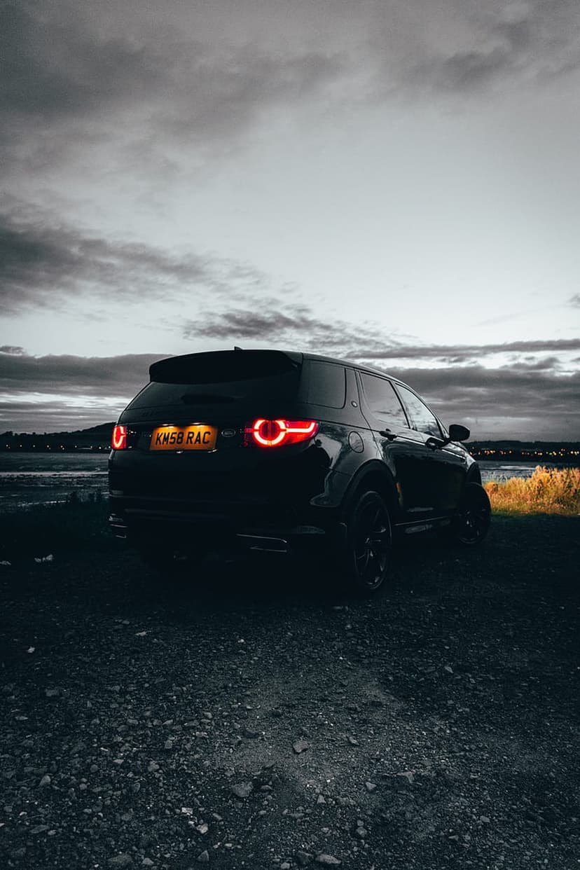 Black SUV with illuminated red taillights parked on gravel near water during a cloudy twilight evening scene.
