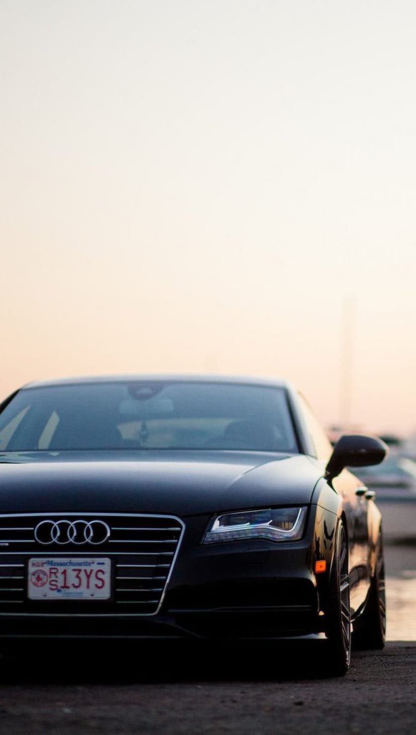 Black Audi A7 front view with glowing LED headlights and a silver grille during a warm outdoor sunset.