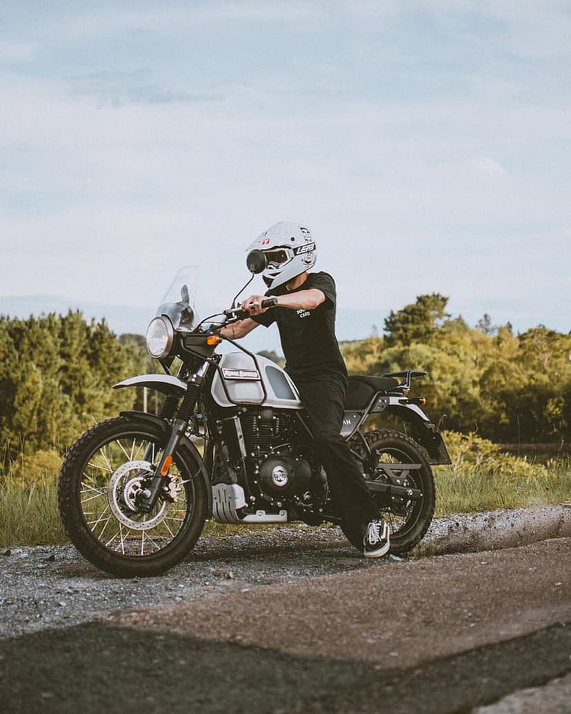 Motorcyclist in a white helmet and black gear sits on a silver bike on a gravel forest road under gray clouds.