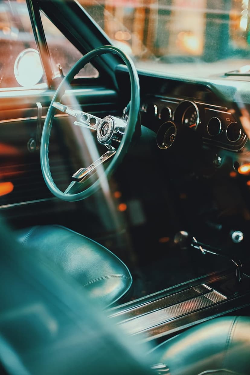Teal leather car seats and a chrome-accented steering wheel sit inside a vintage automobile under warm lighting.