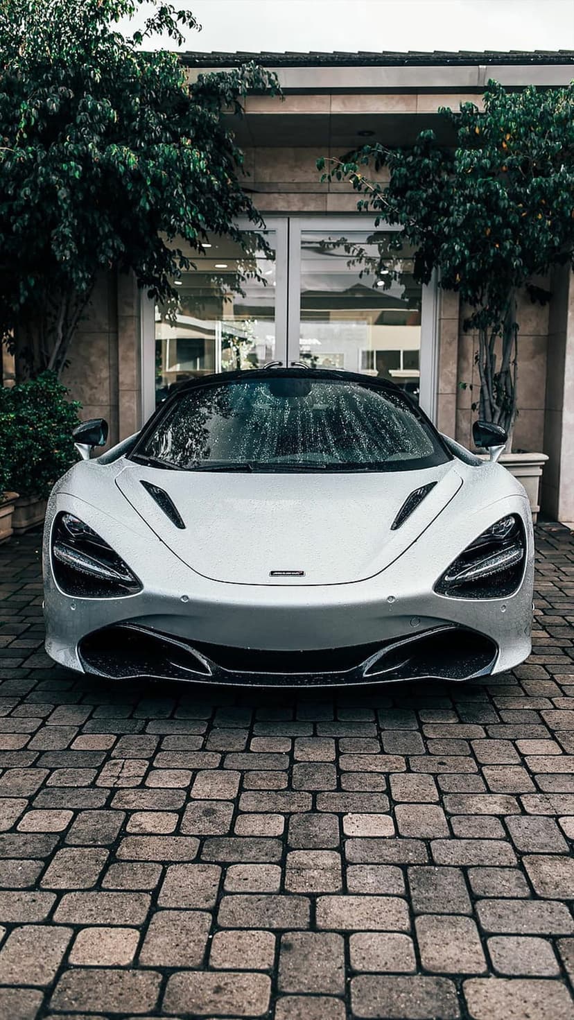 Silver McLaren 720S supercar with raindrops on its hood parked on a cobblestone driveway by a modern building.