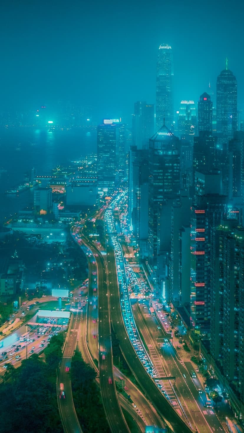 Blue-lit skyscrapers flank a glowing highway with light trails under a misty night sky in a vertical view.
