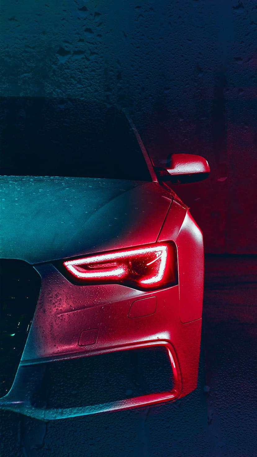 Audi headlights glow with red and blue light on a dark rainy night with visible water droplets on the hood.