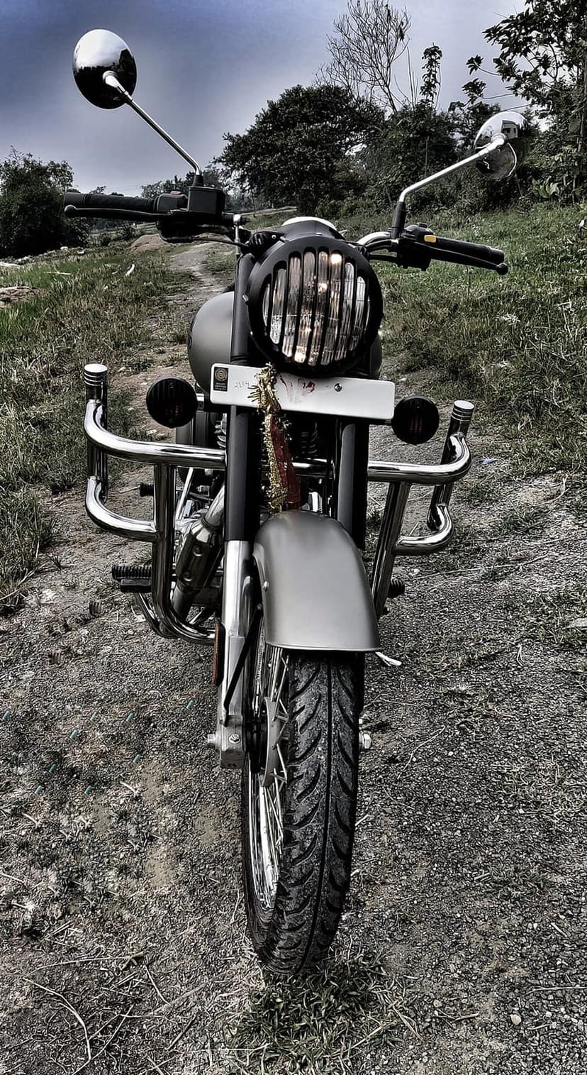 Classic motorcycle front view with a grilled headlight and chrome bars parked on a grassy field under grey clouds.