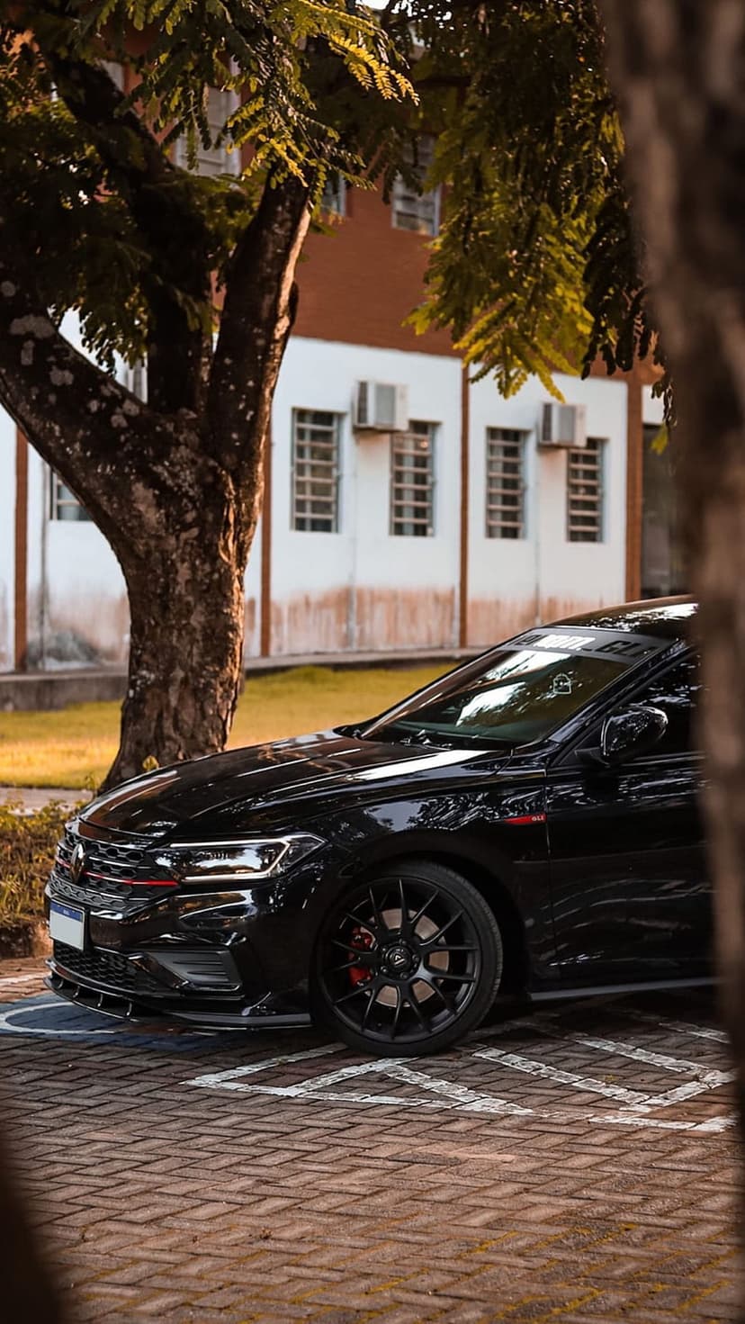 Black sedan with red brake calipers parked on a brick lot under a leafy tree near a modern white building.