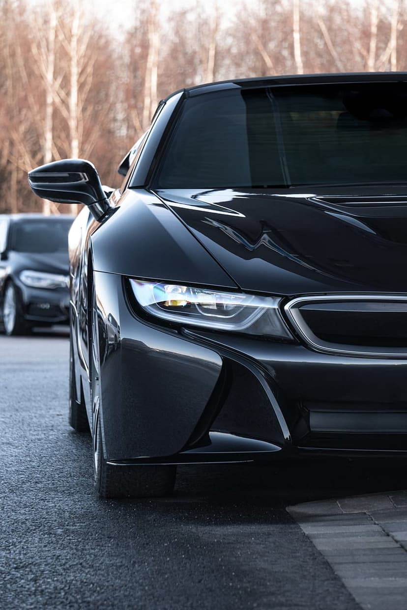 Glossy black supercar front profile featuring illuminated headlights on rain-slicked road with bare trees.