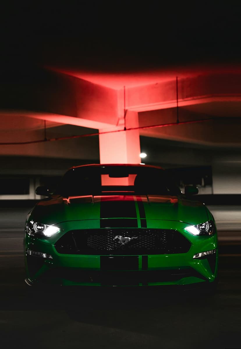 Green Mustang with black stripes sits under red industrial lights with its headlights glowing in the dark.