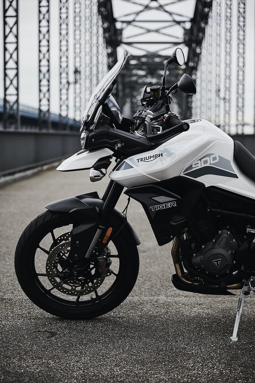 Triumph Tiger 900 motorcycle parked on an asphalt bridge road under a cloudy sky with steel girders.