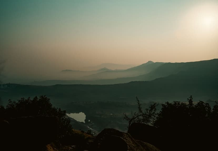 Misty mountain peaks and a still valley lake glow under a soft sunrise framed by dark tree silhouettes.