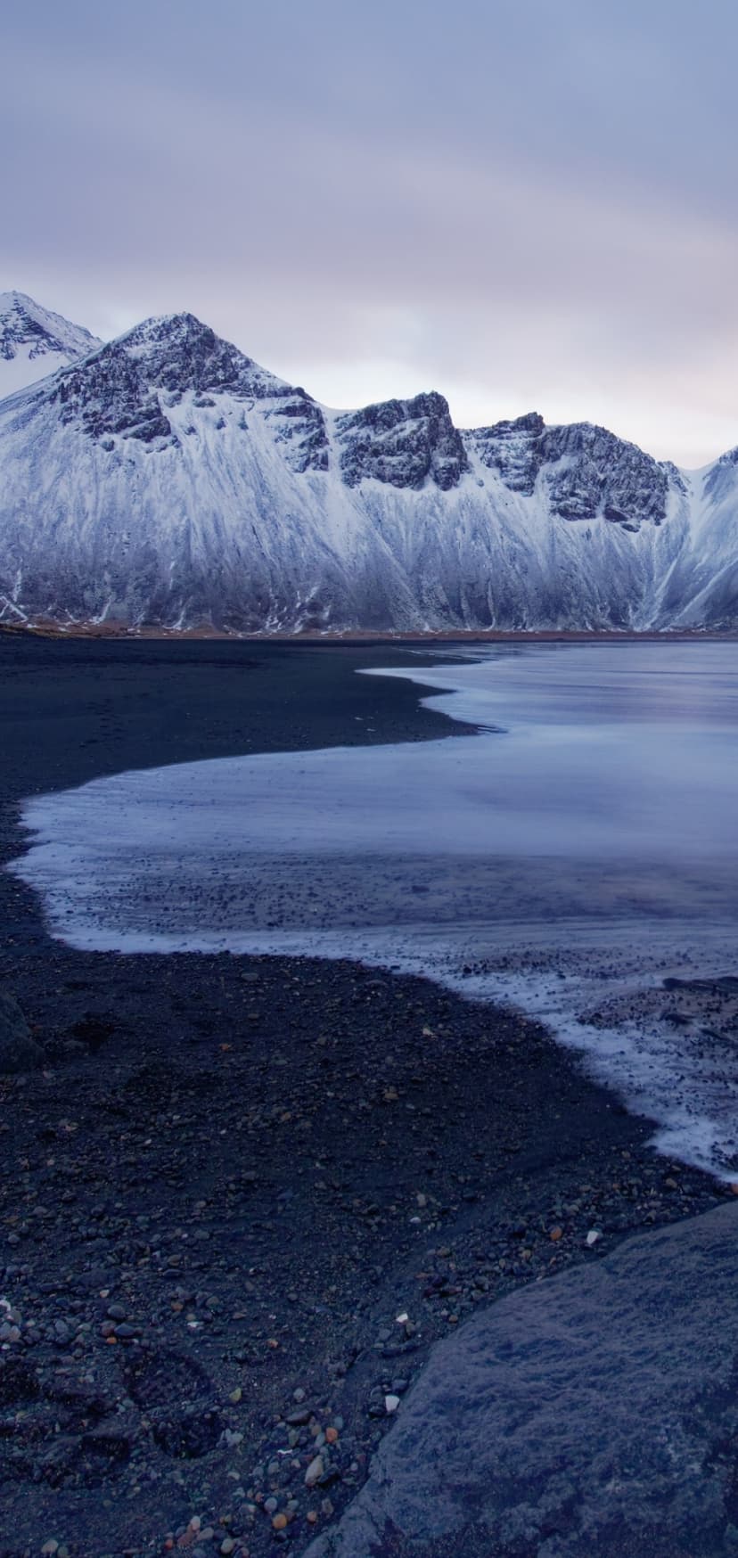 Snow-covered mountains rise above a dark volcanic beach and partially frozen glacial water under a grey sky.