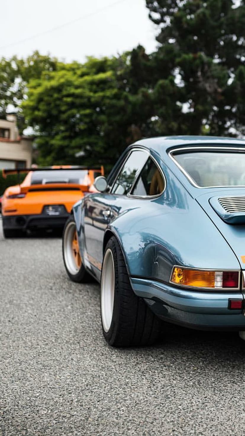Blue Porsche 911 rear view parked on asphalt with an orange sports car and green trees in the background.
