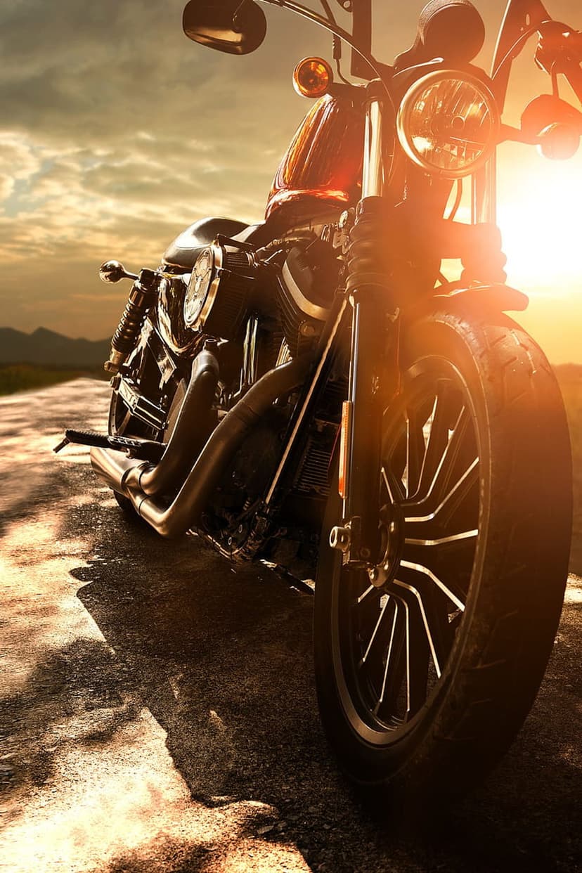 Cruiser motorcycle parked on an asphalt road at sunset with mountain peaks and orange clouds in the background.
